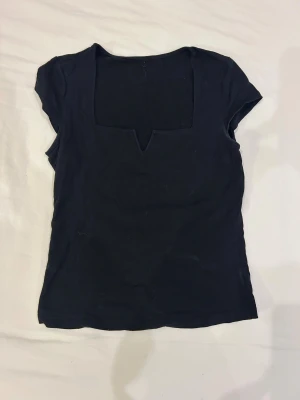 Svart topp med fyrkantig v-ringad hals - Svart topp med korta ärmar och en unik fyrkantig v-ringad hals. Toppens stil är enkel men ändå edgy, perfekt för att lyfta en outfit. Materialet känns mjukt och lätt. 