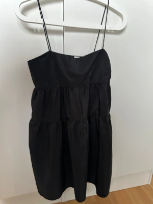 Svart kort klänning från H&M Divided - Supersnygg svart klänning med tunna axelband från H&M Divided. Modellen är kort med voluminösa lager och har en diskret dragkedja i sidan. Materialet är lätt och har en lite glansig finish som ger en cool vibe. Perfekt för dig som gillar minimalistisk stil.