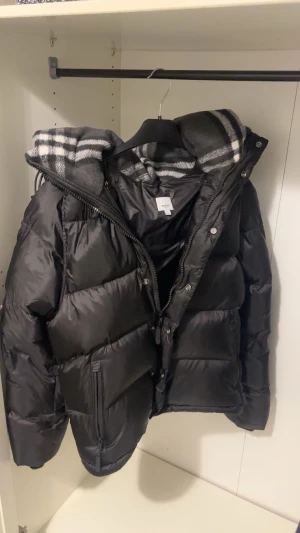 Svart pufferjacka från Burberry - Svart pufferjacka från Burberry med stor huva och klassiskt rutigt foder i grått och vitt. Jackan har dragkedja, knappar och en tydlig Burberry London England-logga på ärmen. Perfekt för kalla dagar med sin tjocka, quiltade design. 