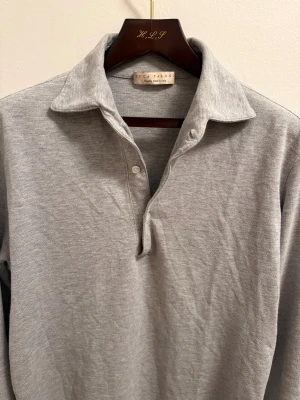 Luca Faloni polo - Snygg grå långärmad pikétröja från Luca Faloni med klassisk krage och två knappar vid halsen. Tröjan är tillverkad i mjuk bomull och har knappar även vid ärmsluten för en clean look. Perfekt för dig som gillar stilrena och enkla plagg.