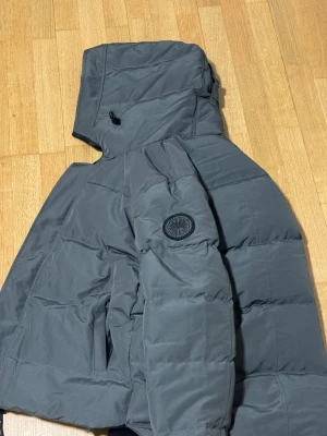  Canada Goose  - Säljer en mörkgrå pufferjacka från Canada Goose med huva och snyggt märke på ärmen. Jackan har dragkedja, praktiska fickor och är fodrad för extra värme. Perfekt för kalla dagar och har en clean, modern look som funkar till streetwear.