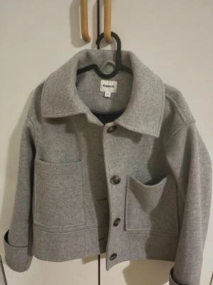 Grå kappa från Kappahl XS - Grå kappa från Kappahl i storlek XS. Jackan har  krage, stora framfickor och knäppning med knappar. Materialet är mjukt och känns som ull, perfekt för lager på lager.🩷🩷🩷Jätte fint skick köpt för 499 kr