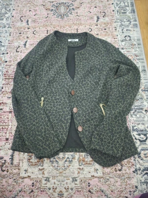 Mönstrad cardigan jacka  - Mörkgrön cardigan jacka med svart triangulärt mönster. Jackan har tre stora bruna knappar framtill och guldfärgade dragkedjor på fickorna. Materialet känns mjukt och är långärmat.