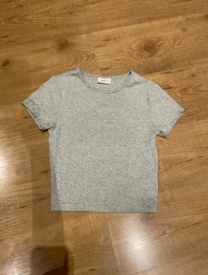 Grå basic t-shirt från Gina Tricot - Enkel och stilren grå t-shirt från Gina Tricot, basically basics-kollektionen. T-shirten har korta ärmar, rund halsringning och är tillverkad i mjuk bomull. Perfekt till jeans eller kjol för en clean look.