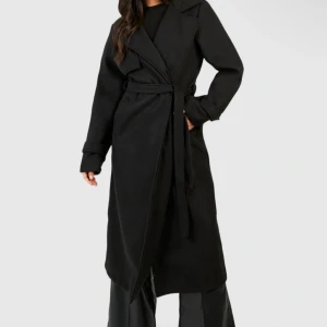 Svart lång kappa från Boohoo - Säljer en svart lång kappa från Boohoo med klassisk krage och bälte i midjan. Kappan har en snygg omlott-design och långa ärmar. Priset går att diskutera vid snabb affär. Endast använd fåtal gånger