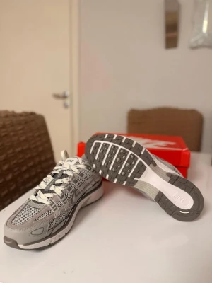 Nike p6000 grå sneakers - Nike p6000 sneakers i grått och vitt med mesh och syntetpaneler. Skorna har en chunky sula, reflekterande detaljer och klassisk Nike Swoosh i silver. Perfekta för dig som gillar sportig och modern stil. Skön passform och snygg design.