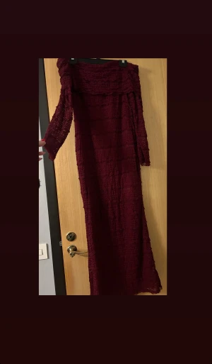 Vinröd långklänning med struktur - Aldrig använd, Vinröd långklänning med snyggt strukturerat tyg och offshoulder-design. Klänningen har långa ärmar och ett elegant, figurnära fall. Perfekt för dig som vill sticka ut med en djup färg och unik textur. Materialet känns lätt och luftigt.