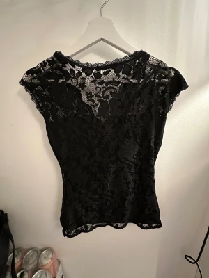 Svart spetsblus från Gina Tricot - Säljer en svart blus från Gina Tricot i storlek S. Toppen är helt i spets med blommigt mönster, v-ringad hals och ärmlös design. Den har en slim passform och transparenta detaljer som ger en snygg look. Perfekt för dig som gillar stilrena och trendiga plagg.
