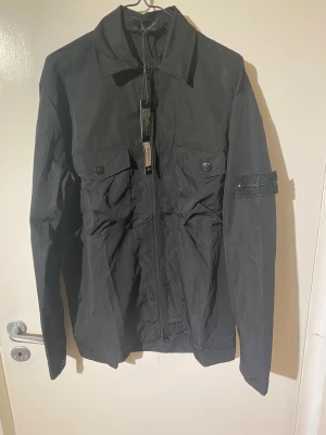 Svart overshirt från Stone Island - Svart overshirt från Stone Island med klassisk krage, två bröstfickor med knappar och ikonisk patch på vänster ärm. Jackan har dragkedja framtill och är tillverkad i lätt polyester. Perfekt för dig som gillar stilrena och funktionella plagg.
