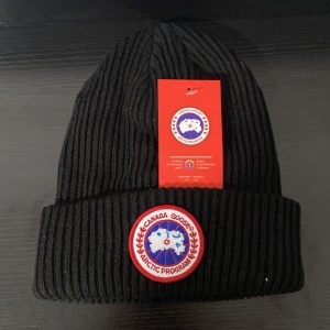 Svart ribbad mössa  - Svart ribbstickad mössa med klassisk rund patch framtill i vitt, rött och blått. Mössan har uppvikt kant och är tillverkad i mjukt stickat material som håller dig varm. Perfekt för kalla dagar och har en clean, tidlös look.