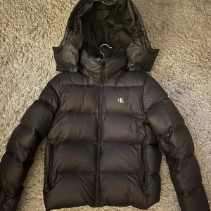 Svart pufferjacka från Calvin Klein - Svart pufferjacka från Calvin Klein med avtagbar huva och diskret CK-logga på bröstet. Jackan har dragkedja framtill och två sidofickor med dragkedja. Den är vadderad och har en snygg, lite boxig passform som är perfekt för kalla dagar. Använd fåtal gånger under förra vinter och säljer då den inte används längre. Ny pris 2600. Pris kan diskuteras!
