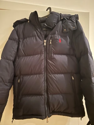 Svart pufferjacka från Polo Ralph Lauren - Svart pufferjacka från Polo Ralph Lauren med röd broderad logga på bröstet. Jackan har huva, flera dragkedjeförsedda fickor och RL-detaljer på dragkedjorna. Perfekt för kalla dagar och riktigt snygg streetstil.