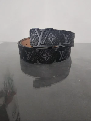 Louis Vuitton svart skinnbälte - Snyggt svart bälte från Louis Vuitton med klassiskt monogrammönster och stor LV-logga i metall på spännet. Bältet är gjort i exklusivt skinn och har en stilren, mörk look som lyfter vilken outfit som helst.