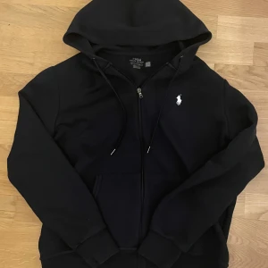 Mörkblå hoodie med dragkedja från Polo Ralph Lauren - Svart hoodie från Polo Ralph Lauren med klassisk vit logga på bröstet. Tröjan har huva med snörning, dragkedja framtill och en stor ficka på magen. Storlek L men passar också bra som storlek M