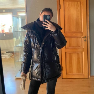 Svart dunjacka Michael Kors XS - Säljer en svart, glansig dunjacka från Michael Kors i storlek XS. Jackan har hög krage, dragkedja framtill och två fickor med dragkedja. Stort MK-märke på ärmen och en oversized look. Jackan har även en luva som man kan ta ut!
