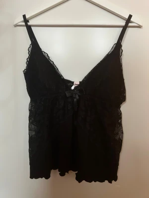 Svart spetsig linnetopp med rosett - Säljer en svart linnetopp med tunna axelband och v-ringad spets framtill. Toppen har en söt liten rosett i satin mitt på bröstet och är dekorerad med blommig spets längs kanterna. Perfekt för dig som gillar romantiska detaljer och en feminin look.