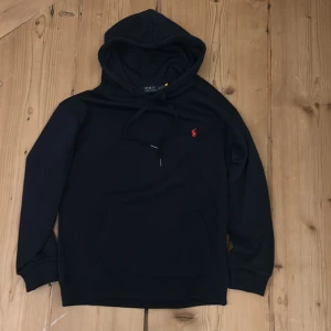 Svart hoodie från Polo Ralph Lauren - Svart hoodie från Polo Ralph Lauren med klassisk röd logobrodyr på bröstet. Tröjan har huva med snörning, stor magficka och ribbade muddar vid ärmslut och nederkant. Perfekt för chill dagar och streetwear-stil.