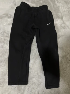 Svarta mjukisbyxor från Nike, M - Svarta mjukisbyxor från Nike i storlek M med klassisk vit swoosh-logga på benet. Byxorna har elastisk midja och är gjorda i mjukt bomullsmaterial, perfekta för chill eller träning. Snygg och enkel design som funkar till sneakers.       81 cm lång 43 cm bred