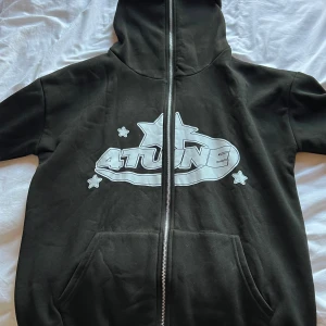 Svart hoodie med 4tune-tryck - Svart hoodie från 4tune med stor vit logga och stjärnor på framsidan samt cirkulärt tryck med 4tune-text och stjärnor på ryggen. Tröjan har huva, dragkedja och rymliga fickor framtill. Perfekt för dig som gillar streetwear och grafiska prints.