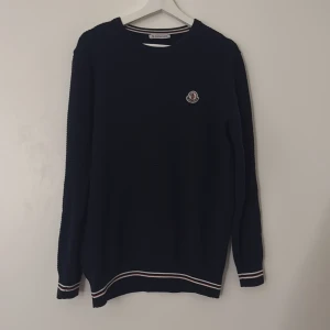 Mörkblå stickad tröja från Moncler - Snygg mörkblå stickad tröja från Moncler med rund hals och diskret logga på bröstet. Tröjan har vita och röda ränder vid muddar och nederkant, vilket ger en chill vibe. Perfekt för dig som gillar stilrena och exklusiva plagg. Pris kan diskuteras ifall man inte är nöjd med det!