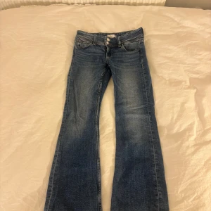 Blå bootcut jeans från Gina Tricot - Snygga blå jeans från Gina Tricot i storlek 146 med klassisk bootcut passform. Jeansen har två framfickor, dubbla knappar i midjan och bakfickor med knappdetalj. Materialet är jeans med lätt tvättad look och kontrastsömmar.