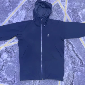 Svart vindjacka från Haglöfs med huva - Säljer en mörkblå vindjacka från Haglöfs i storlek S. Jackan har hel dragkedja framtill, justerbar huva och minimalistisk design med diskret logga på bröstet. Perfekt för blåsiga dagar och outdoor-äventyr.