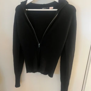Svart hoodie med dragkedja Gina Tricot - Svart hoodie från Gina Tricot i storlek M. Tröjan har ribbad nederdel, lång ärm och huva. Materialet är stickat och den har en dragkedja framtill. Perfekt för dig som gillar enkel och stilren look.