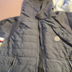 Grå quiltad jacka från Moncler - Snygg grå quiltad jacka från Moncler med huva och dragkedja framtill. Jackan har Moncler-logga på bröstet, två fickor med dragkedja och en stilren design. Perfekt för dig som gillar en clean look och vill ha något som funkar till många outfits. Passar även s