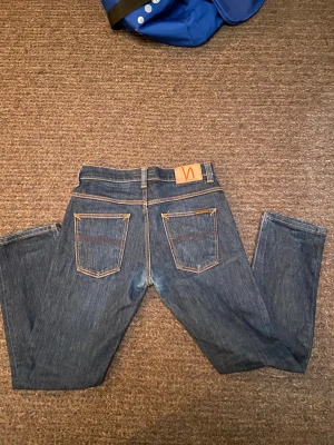 Blå jeans från Nudie Jeans Co W32 L32 - Snygga blå jeans från Nudie Jeans Co i klassisk modell med raka ben och orangea kontrastsömmar. Jeansen har fem fickor och en patch med Nudie-loggan bak. Passar dig som gillar stilrena och tidlösa jeans med schysst passform.