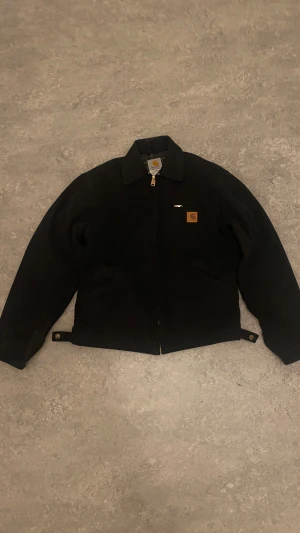 Svart Carhartt bomberjacka - Svart bomberjacka från Carhartt med klassisk krage, dragkedja framtill och brunt logopatch på bröstet. Jackan har två snedställda fickor och en bröstficka med dragkedja. Insidan är fodrad med rutigt tyg för extra komfort. Perfekt för dig som gillar streetwear.