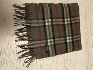 Rutig grå halsduk från Burberry - Snygg halsduk från Burberry i klassiskt rutigt mönster med grå, vit, svart och röda toner. Gjord i 100% kashmir för extra mjuk och lyxig känsla. Långa fransar i ändarna ger en cool touch. Perfekt accessoar för att lyfta din vinterstil.