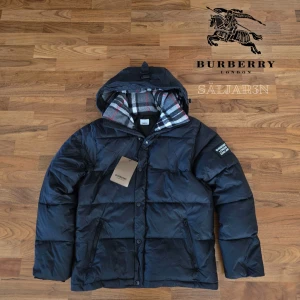 Burberry Puffer  - Svart dunjacka från Burberry med klassisk Säljer en elegant och mångsidig Burberry-jacka i svart, i nyskick. Dunfodrad för värme och komfort med det klassiska rutmönstret i huvan som ger en exklusiv touch. Praktiska dragkedjefickor, avtagbar huva och diskret logopatch på ärmen. Jackan är i princip som ny och utan defekter. En lyxig kombination av stil och funktion – perfekt för både vardag och tillfällen då du vill klä upp dig.  📦Snabb leverans - skickas samma dag📦 