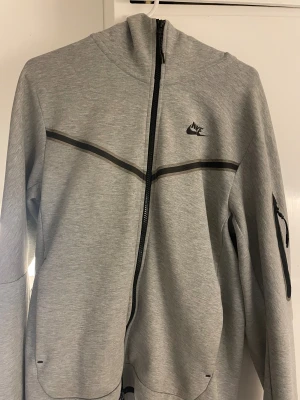 Grå Nike tech ziphoddie - Snygg grå nike tech med svart logga på bröstet och svarta detaljer. Jackan har huva, hel dragkedja framtill och en extra ficka med dragkedja på ärmen. Perfekt för dig som gillar sportig stil och vill ha något bekvämt och funktionellt. 