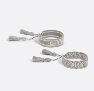 Grå broderade armband från Dior - Två snygga grå armband från Dior med broderad logga och mönster. Armbanden har tvinnade snören och dekorativa tofsar i ändarna. Perfekt accessoar för att ge din outfit en exklusiv touch. Justerbar passform med knytning.