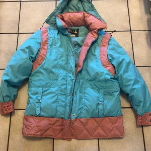 Turkos och rosa pufferjacka med huva - Retro pufferjacka från OE i turkos och rosa med quiltade detaljer och stor huva. Jackan har dragkedja och tryckknappar framtill, två fickor med lock och elastiska muddar vid ärmslut. Fodret är tjockt och jackan har en loose passform, perfekt för kalla dagar.