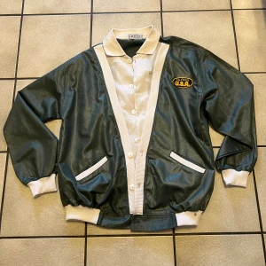 Grön bomberjacka från Hugo Boss  - Retro bomberjacka från Hugo Boss i mörkgrönt med vita ribbade muddar och krage. Jackan har broderad patch med 'California U.S.A.' på bröstet, vita knappar och två fickor fram. Materialet är syntet och jackan har en loose passform.