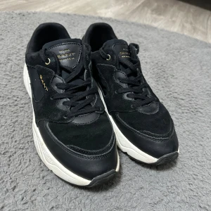 Svarta sneakers Gant i mocka och skinn - Snygga svarta sneakers från Gant med detaljer i mocka och skinn, vit chunky sula och guldfärgade logotyper. Skorna har snörning och en cool kontrast mellan material och färger. Perfekta för dig som gillar stilrena men sportiga skor. Passar även 39,5! Nypris: 1500kr 