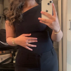 Svart oneshoulder topp med guld detalj  - Snygg svart topp med oneshoulder-design med guldig detalj som ger en cool och modern look. Toppen är tight och har en slät yta utan synliga detaljer eller mönster. Perfekt för dig som gillar stilrena plagg med en twist.