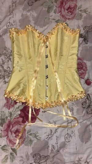 Gul korsett med blommigt mönster och snörning - Säljer en ljusgul korsett med blommigt mönster i glansigt tyg. Den har vackra volangdetaljer upptill och nedtill, samt snörning och satinband både fram och bak. Materialet känns som satin eller polyester och ger en lyxig känsla. Perfekt för dig som vill sticka ut! Använt en gång på halloween, råkat få nån röd färg på korsetten, går kanske att ta bort men har inte testat ännu.