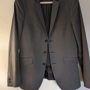 Grå kavaj från HOPE Stockholm - Snygg grå kavaj från HOPE Stockholm med klassisk passform och tre knappar framtill. Kavajen har två fickor med lock och en bröstficka. Materialet är mjukt och har en lätt struktur, perfekt för en stilren look. Fodrad insida med HOPE-logga.