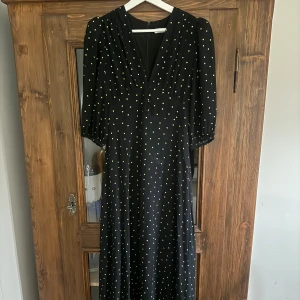 Svart prickig långklänning med v-ringning - Svart långklänning med ljusgula prickar, djup v-ringning och knappar framtill. Klänningen har trekvartsärmar med lätt puff och resår vid ärmslut. OBS! Se bilder för skick, fläckar på framsida (kanske går bort med lätta medel, har inte provat)