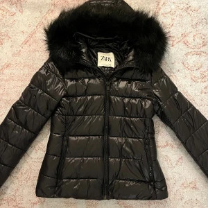 Svart pufferjacka med fuskpäls från Zara - Snygg svart pufferjacka från Zara med glansig finish och huva med fluffig fuskpäls. Jackan har dragkedja framtill, två fickor med dragkedja och ribbade muddar vid ärmsluten. Perfekt för kalla dagar när du vill hålla stilen.