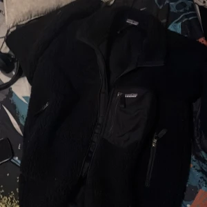 Svart teddyjacka från Patagonia L - Mysig svart teddyjacka från Patagonia i storlek L. Jackan har hög krage, flera dragkedjeförsedda fickor och är tillverkad i mjuk fleece. Perfekt för kyliga dagar och har en clean, stilren look med diskret logga på bröstet.