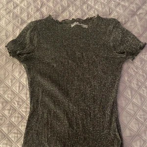 Glittrig svart topp från NA-KD - Säljer en svart glittrig topp från NA-KD i storlek XS💐💕 Toppen har korta puffärmar och en söt volangkant vid halsen och ärmsluten🌸🌷 Materialet är stretchigt och skimrar snyggt i ljuset, perfekt för att sticka ut. Supersnygg att styla med jeans eller kjol!☀️🌻