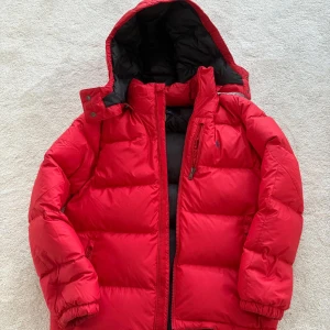 Polo Ralph Lauren jacka st 14-16/160cm - Riktigt fet röd dunjacka från Polo Ralph Lauren med avtagbar huva, vattenavvisande ripstop-tyg och dragkedjor. Jackan har svarta detaljer på insidan och flera fickor med dragkedja. Perfekt för kalla dagar när du vill hålla stilen och värmen.