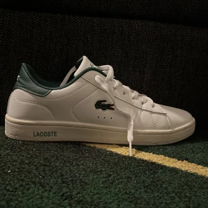Vita sneakers från Lacoste - Snygga vita sneakers från Lacoste med gröna detaljer och klassisk krokodil-logga på sidan. Skorna har snörning, rund tå och är tillverkade i skinnliknande material. Perfekta för en clean och sportig stil.