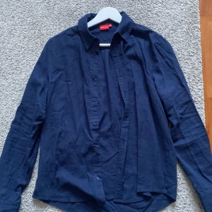 Mörkblå skjorta från RYVLS - Snygg mörkblå skjorta från RYVLS i storlek 158/164. Skjortan har klassisk krage, knappar framtill och långa ärmar. Tillverkad i mjukt linnematerial som känns chill och skönt mot huden. Perfekt för dig som gillar stilrena och enkla plagg.