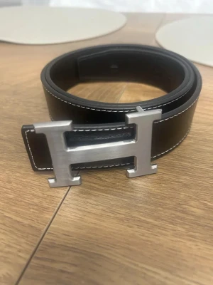 H3rmes bälte - 🖤 Svart bälte med silver H-spänne – 110 cm  Elegant och tidlöst bälte med ett kraftigt H-format spänne i silverfärg. Svart läderlook med vit söm som ger en stilren kontrast. Passar perfekt till både jeans och finare byxor.  📏 Längd: 110 cm 💼 Passar både vardag och fest 🚚 Fraktas och skickas snabbt! 💰 Pris: 279 kr