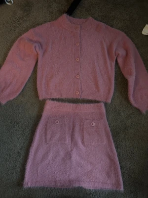 Ljusrosa set - Supermysig ljusrosa set i fluffigt stickat material. Kjolen har två framfickor med matchande rosa knappar som detalj. Koftan har uppknäppbara knappar. Perfekt för dig som gillar mjuka och söta plagg med lite extra cozy vibe. (lite större i storleken)