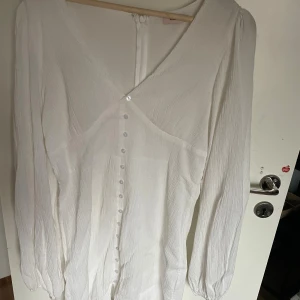 Vit kort klänning från Nelly - Supersöt vit kort klänning från Nelly med v-ringning, långa puffiga ärmar och knappar framtill. Klänningen har en lätt skrynklig struktur och är figurnära upptill. Perfekt för dig som vill ha en fräsch och stilren look.